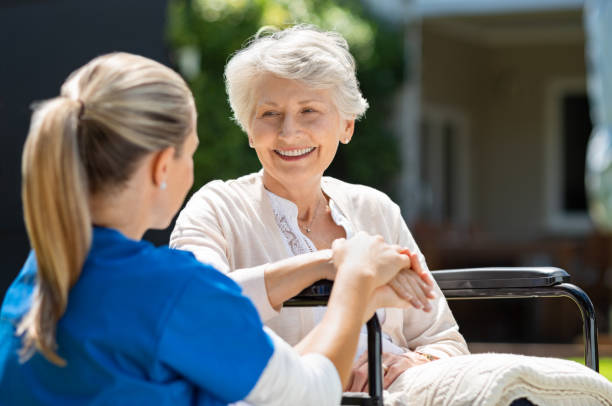 Smiling senior patient sitting in a wheelchair with nurse supporting her. Doctor looking at an elderly patient in a wheelchair in the garden. Nurse holding the hand of a mature woman outside the pension home.