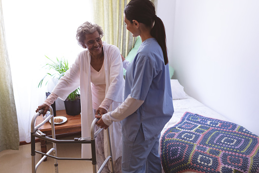 Front view of mixed race female nurse helping senior African American female patient to stand with a walker at a nursing home