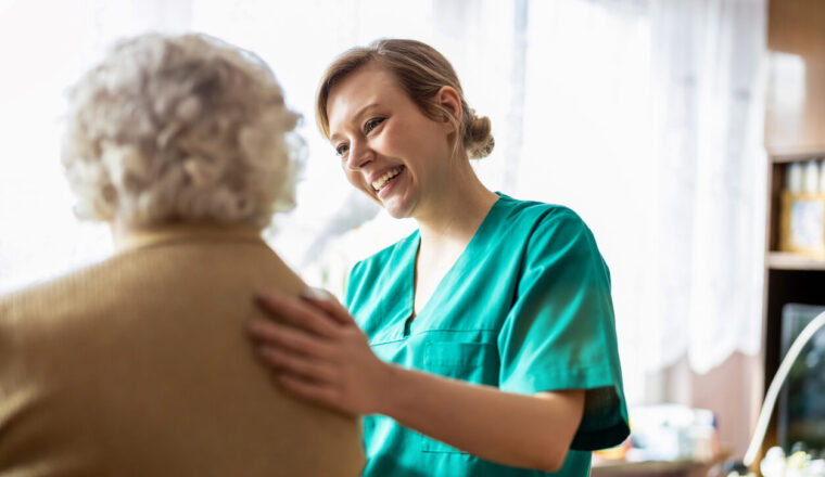 A cheerful caregiver in green scrubs comforts an elderly person, placing a supportive hand on their shoulder in a bright room