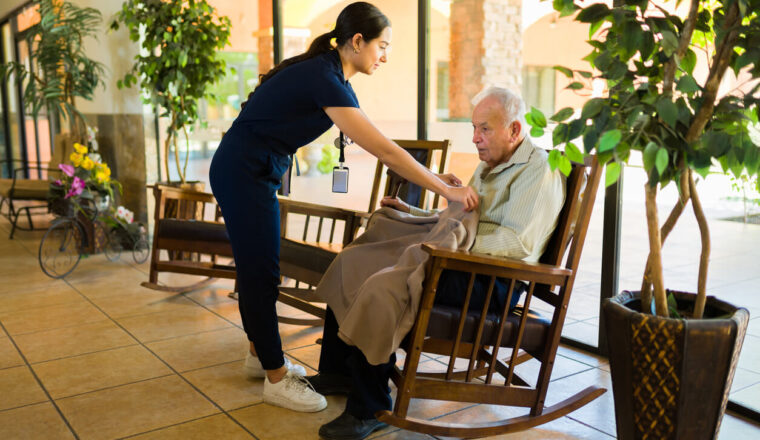 Caring female nurse working at the nursing home giving a warm blanket to an old man sitting on a rocking chair
