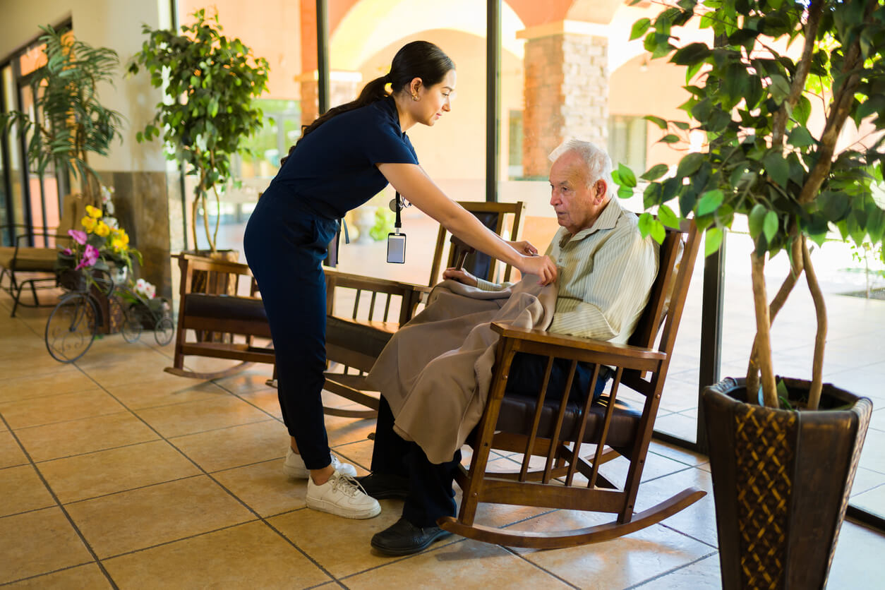 Caring female nurse working at the nursing home giving a warm blanket to an old man sitting on a rocking chair