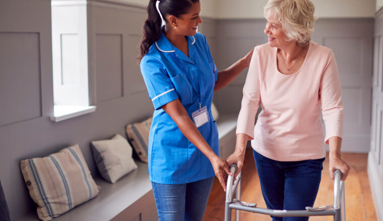 Senior Woman At Home Using Walking Frame Being Helped By Female Care Worker In Uniform
